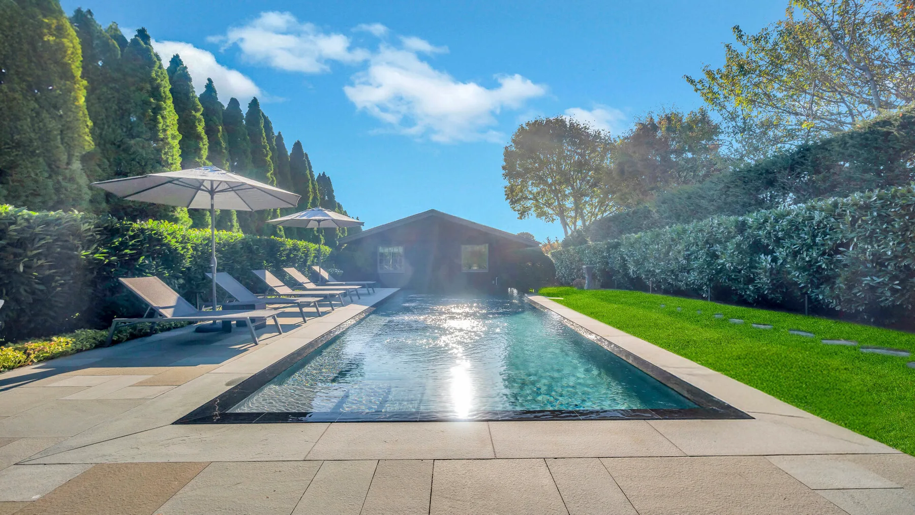An outdoor swimming pool beside a paved terrace, with loungers and greenery.