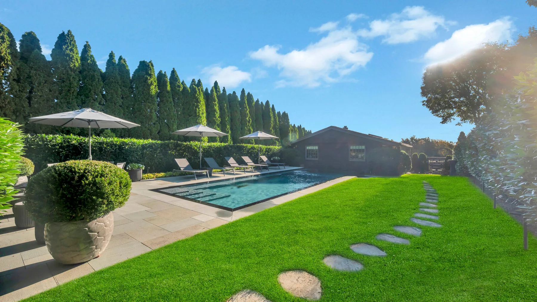An outdoor swimming pool sits beside a lawn, with stepping stones and umbrellas.