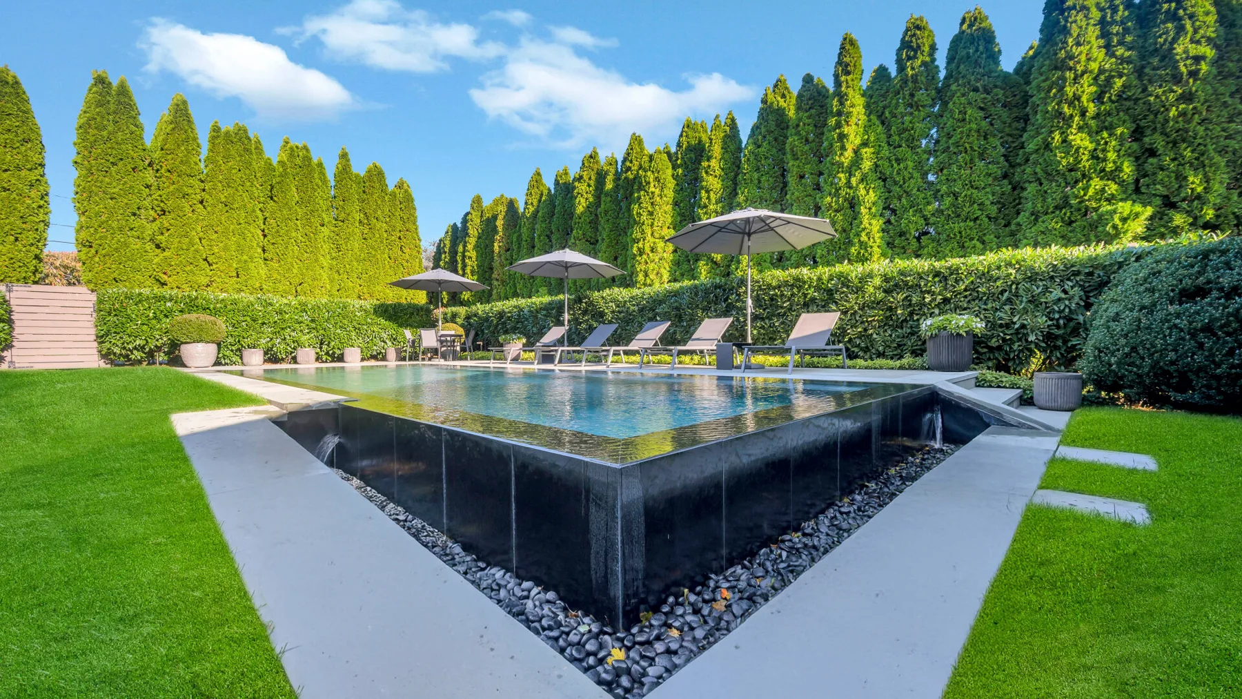 An outdoor swimming pool is surrounded by lawn, hedges, and lounge seating.