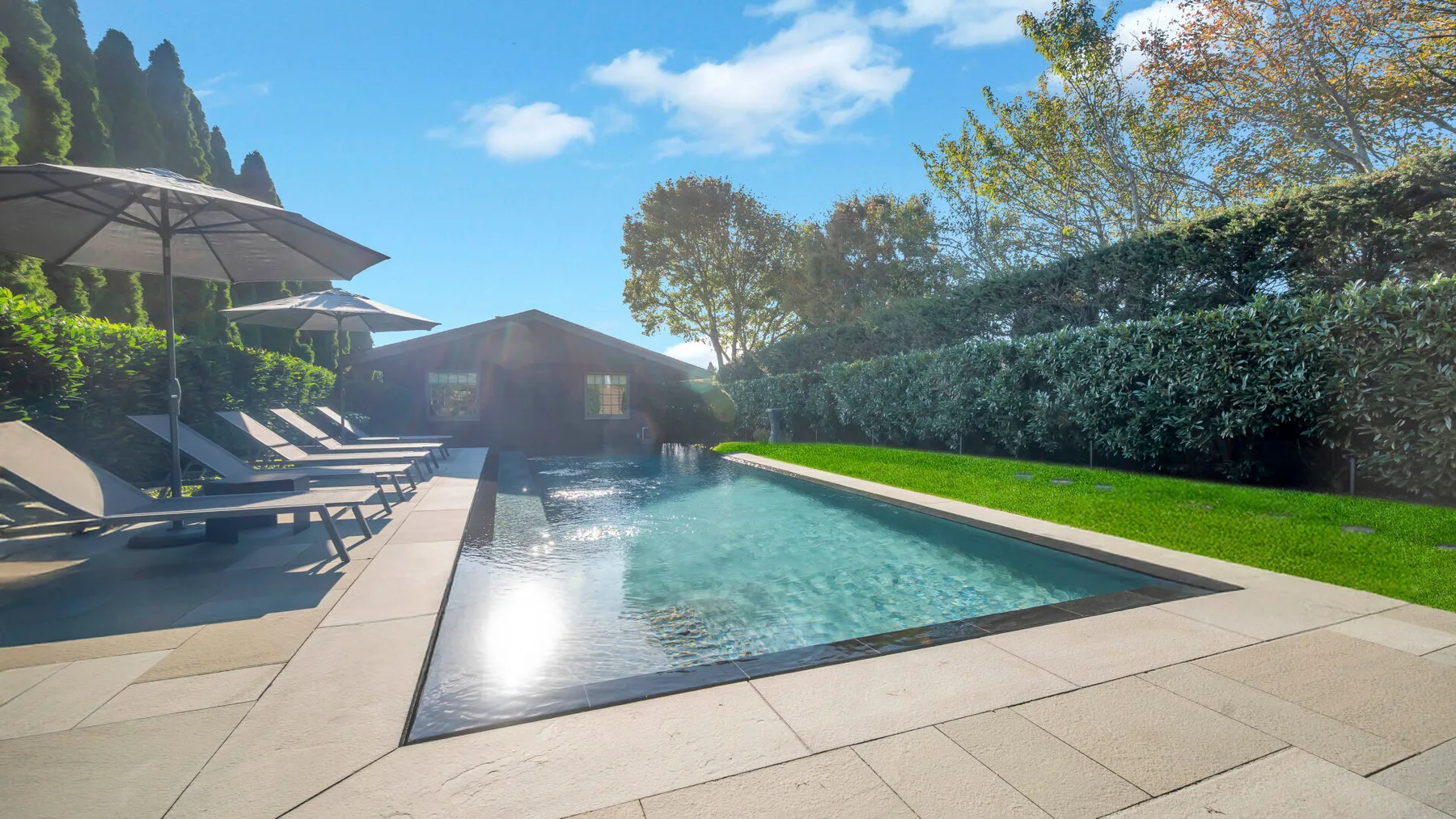 An outdoor swimming pool beside a paved patio with lounge seating and umbrellas.