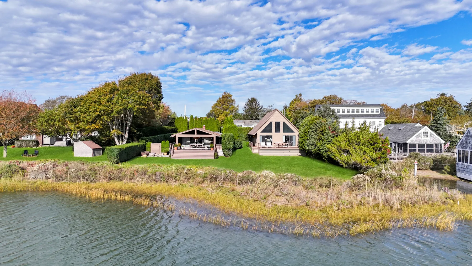 A row of houses and garden buildings sits beside a grassy shoreline.