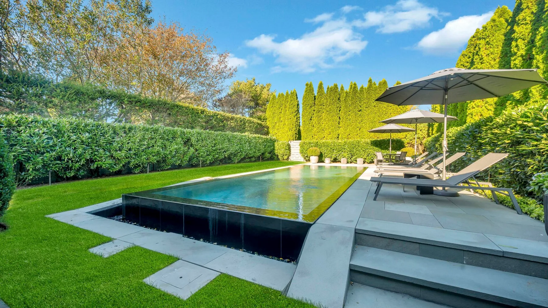 An outdoor swimming pool beside a paved patio, lounge seating, and greenery.
