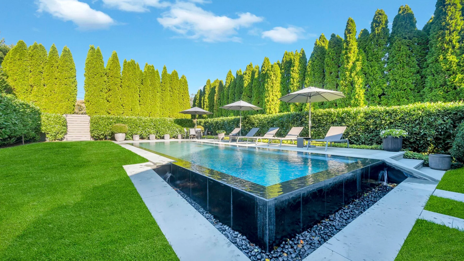 An outdoor swimming pool sits beside a green lawn and tall hedges.