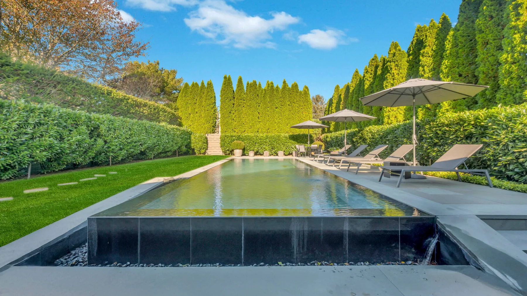 An outdoor pool sits beside a lawn, with lounge chairs and umbrellas along the hedge.