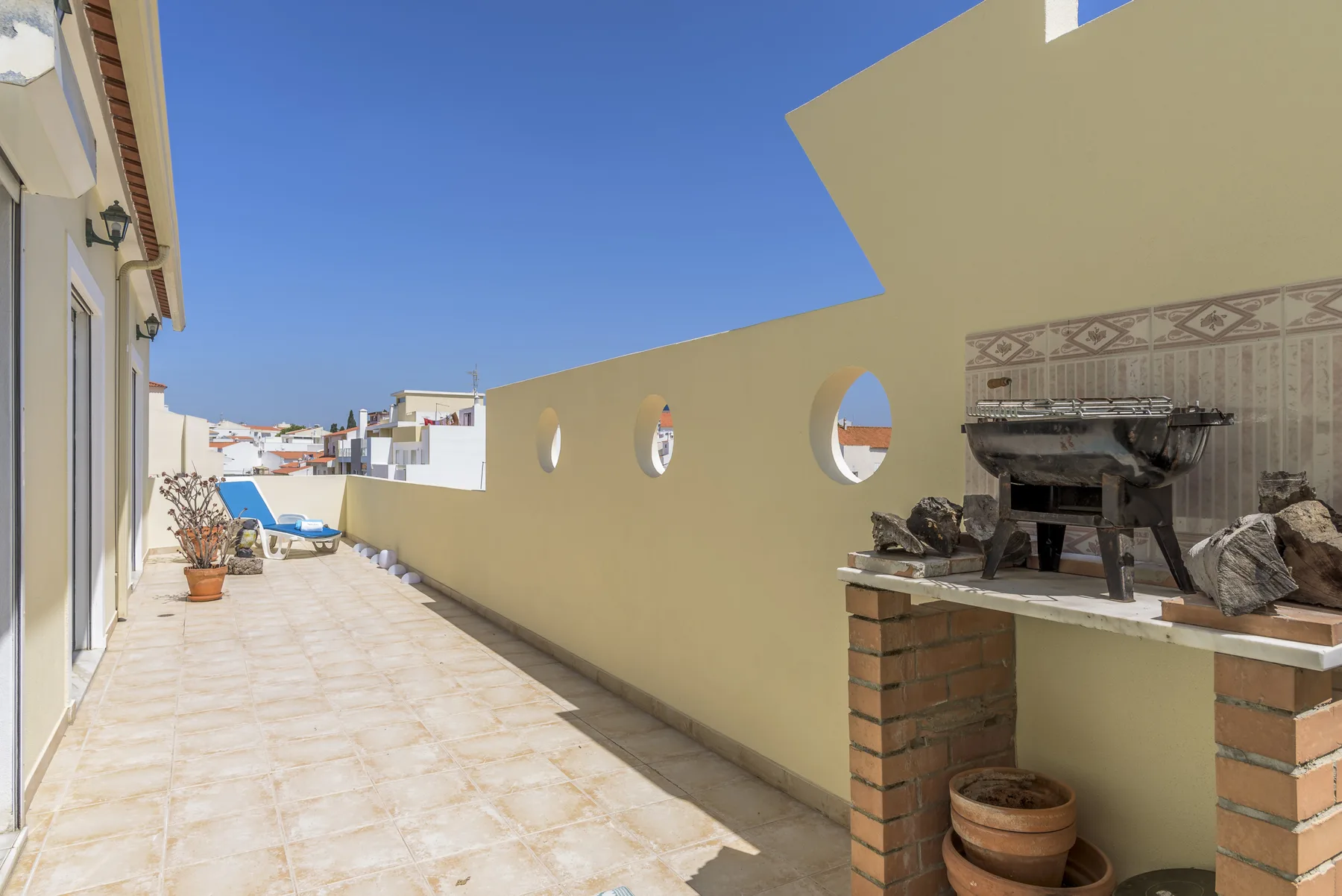 Una terraza soleada en la azotea tiene suelo de baldosas, un muro bajo y una parrilla exterior.