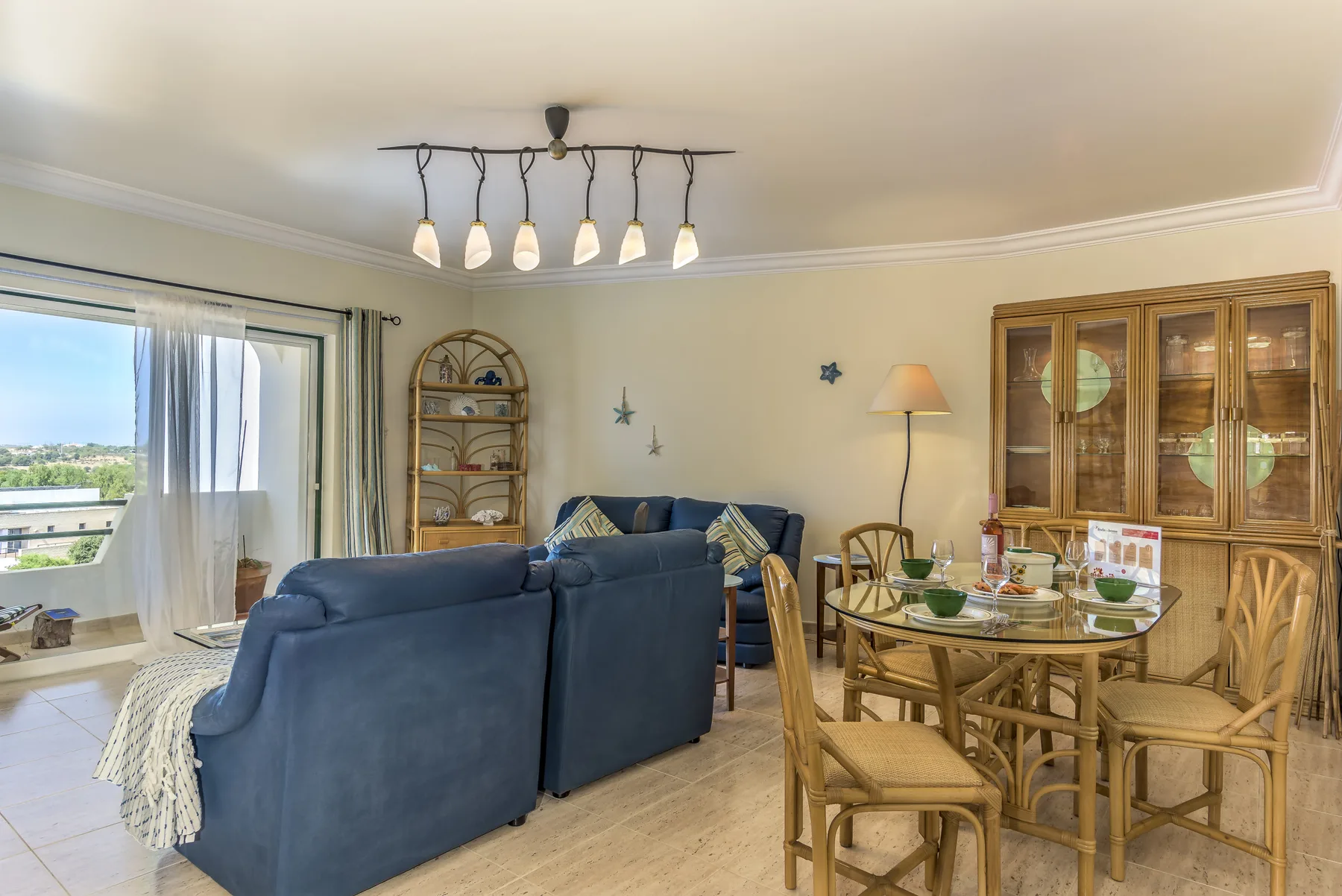 Zona de estar y comedor con asientos azules, una mesa de madera y ventanales.