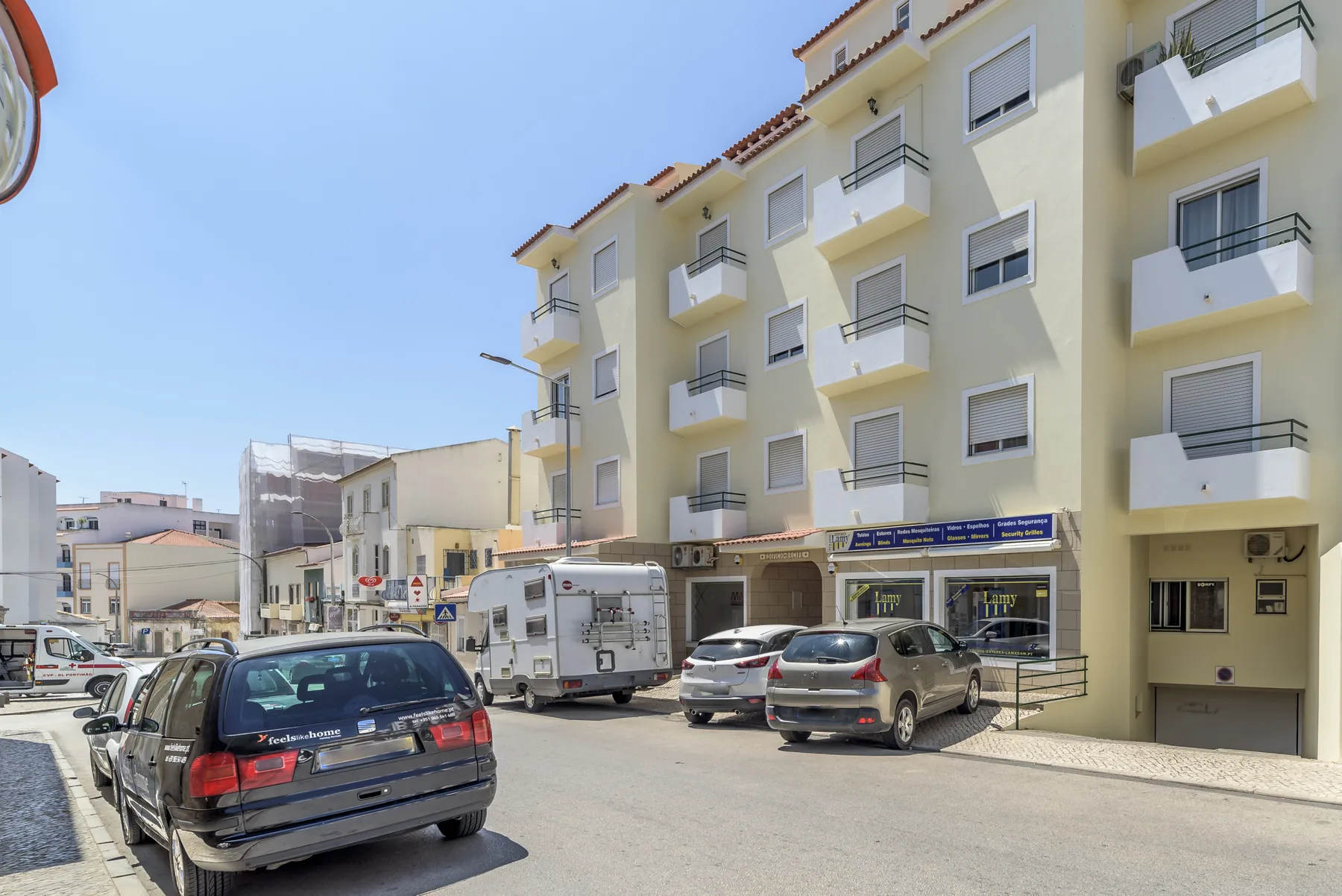 Una calle soleada muestra un edificio de apartamentos claro con balcones y coches estacionados.