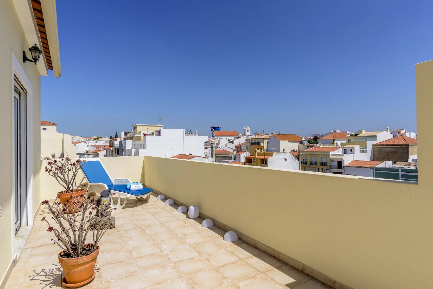 Una terraza soleada en la azotea con plantas en macetas y vistas de edificios cercanos.