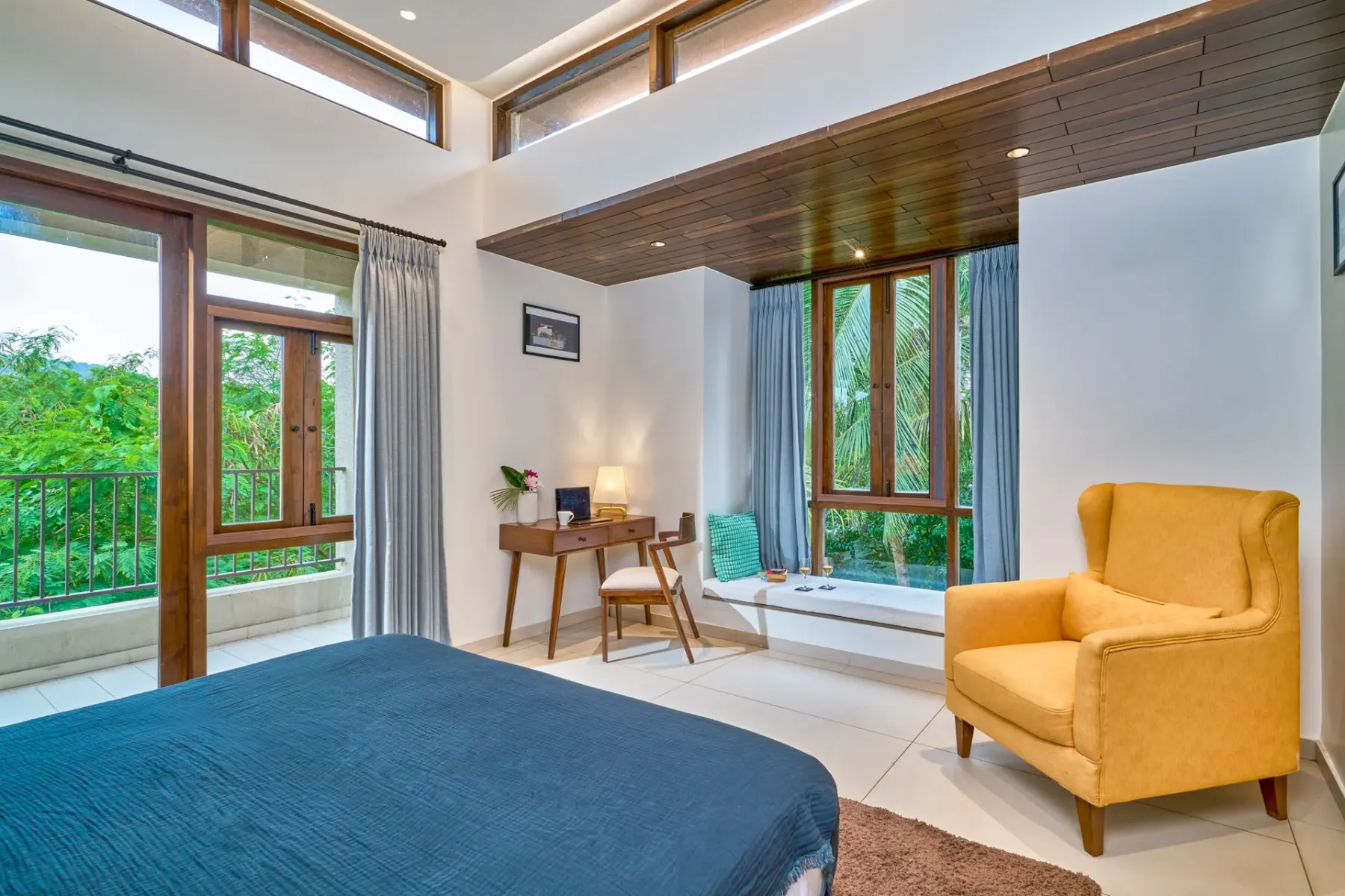 A bright bedroom with a blue bed, desk, and mustard-colored armchair.
