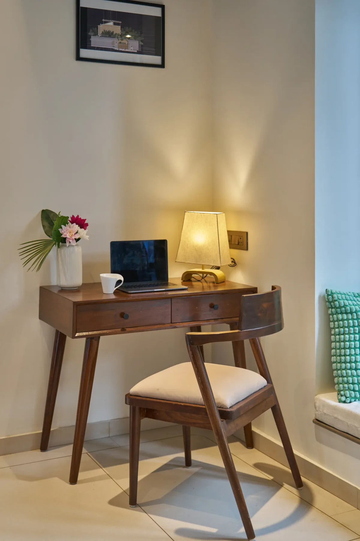 A desk and chair sit beside a window, with a lamp and flowers on the desk.