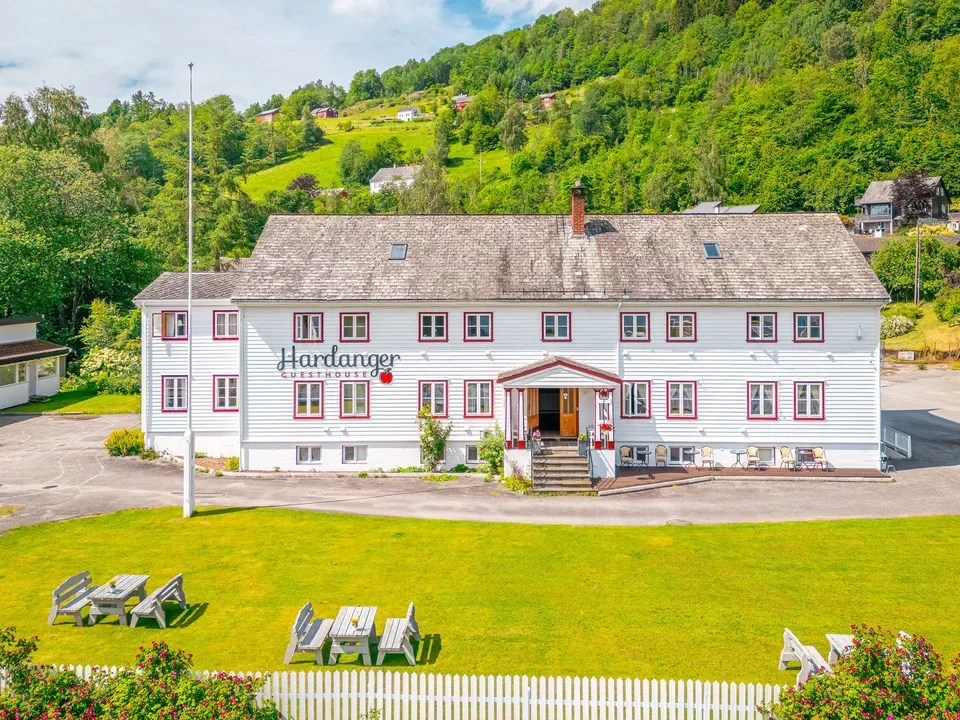 White guesthouse with red trim on a green lawn, backed by a wooded hillside.