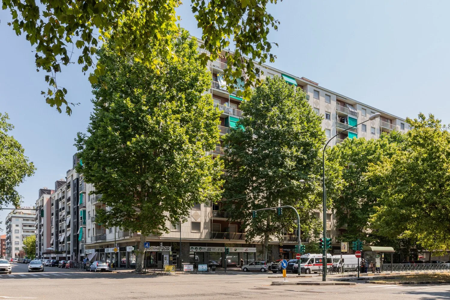 A city street corner with a mid-rise building partly screened by leafy trees.