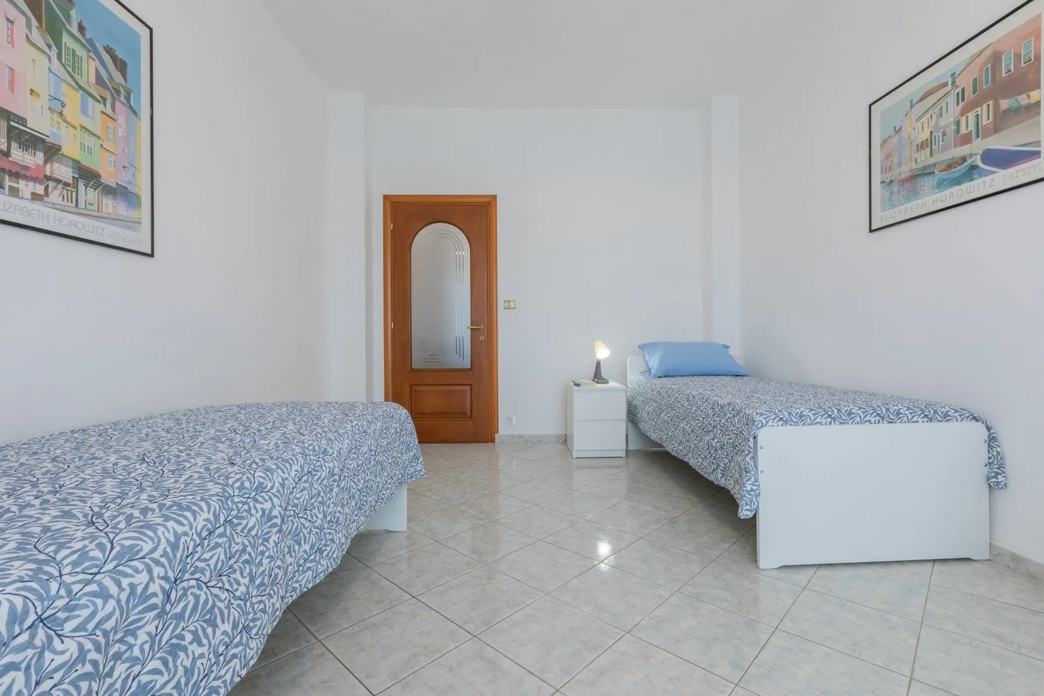 A bright bedroom with two single beds and framed artwork on the walls.