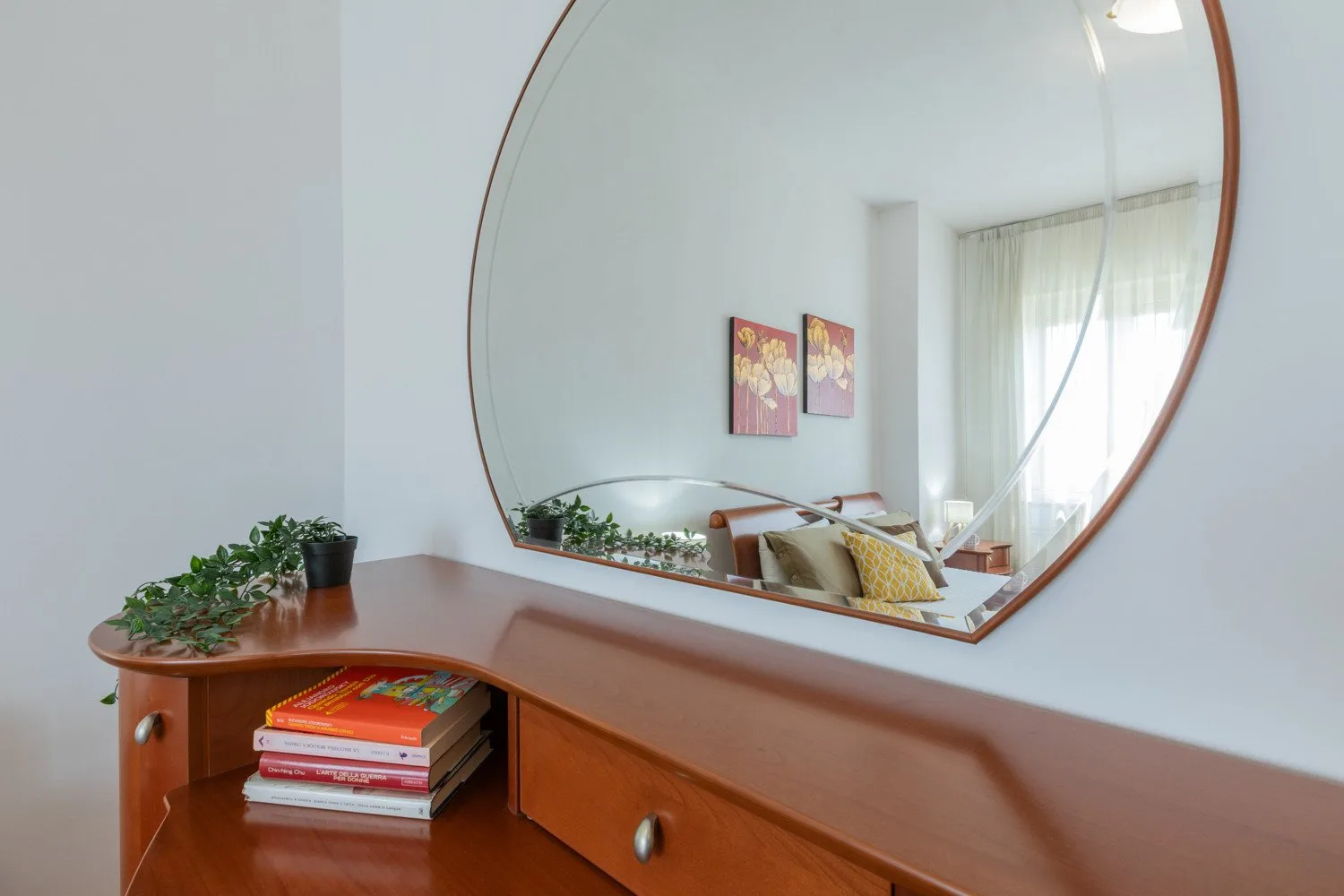 A wooden dresser with books and plants sits beneath a large round mirror.