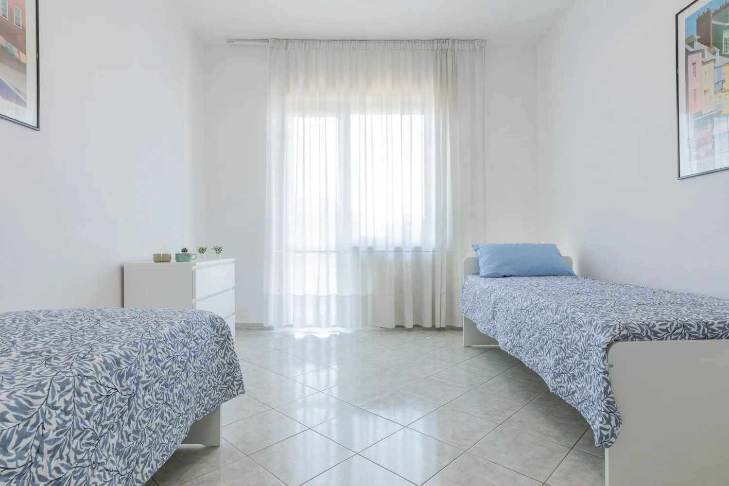A bright bedroom with two beds and a curtained window.