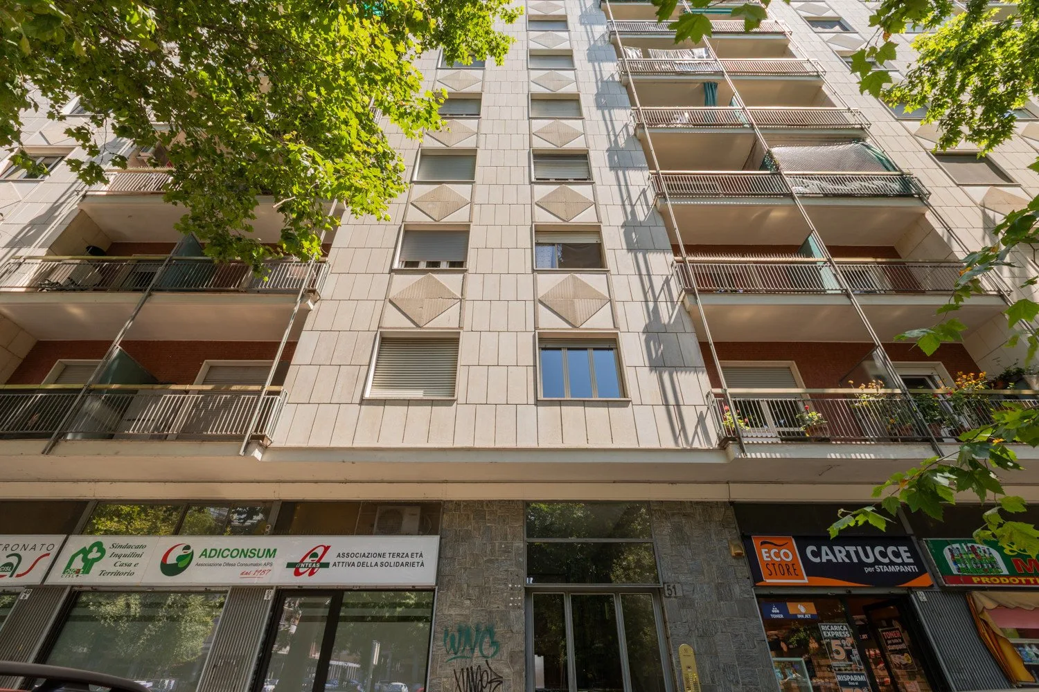 An apartment building viewed from below, with balconies and leafy trees.