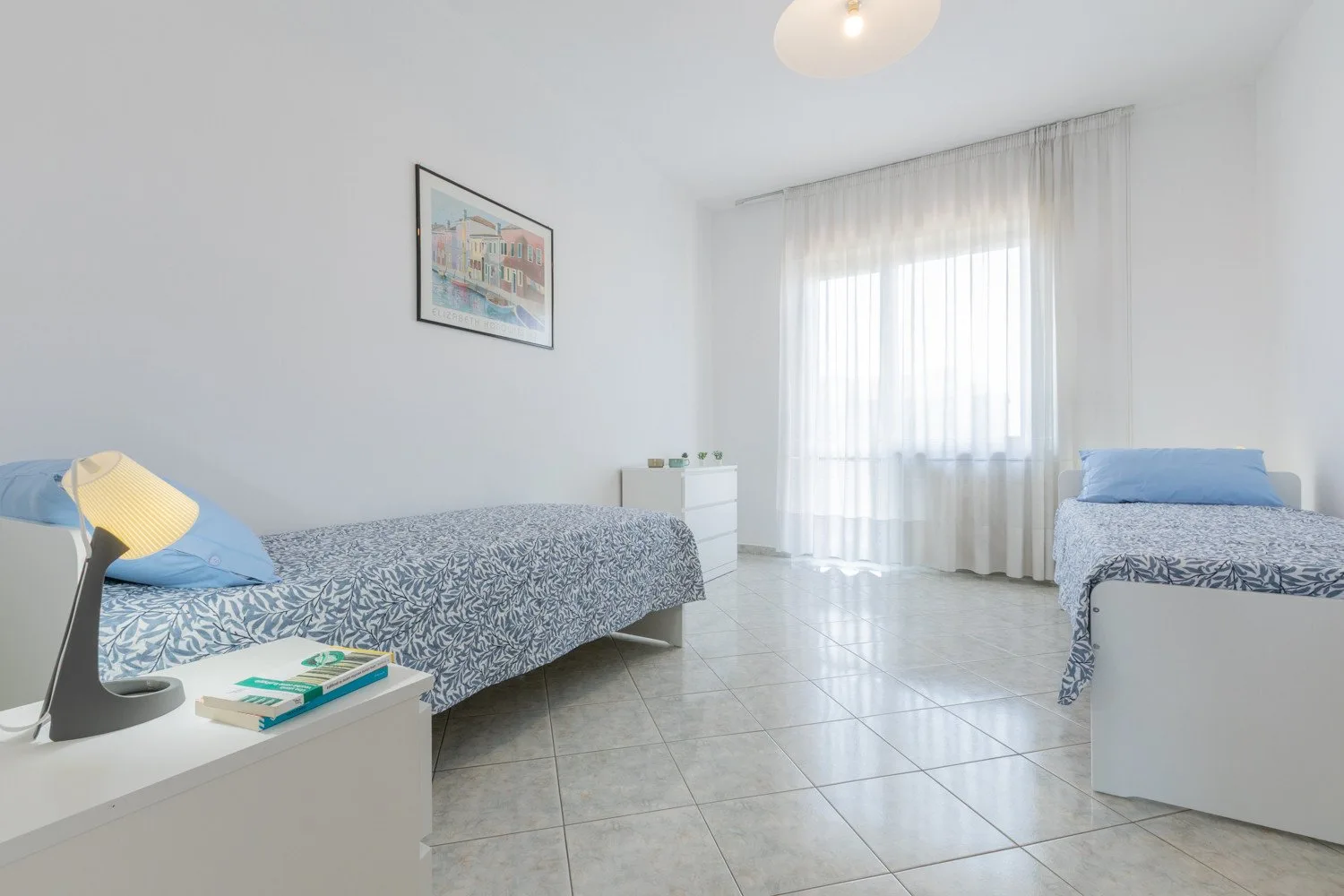 A bright bedroom with two beds and sheer curtains over a large window.