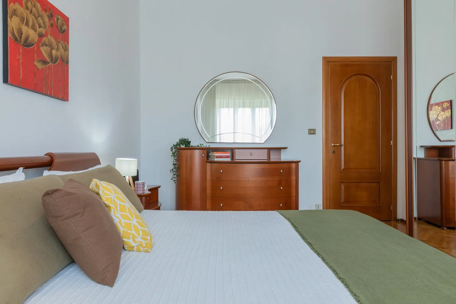 A bedroom with a neatly made bed and a wooden dresser beneath a round mirror.