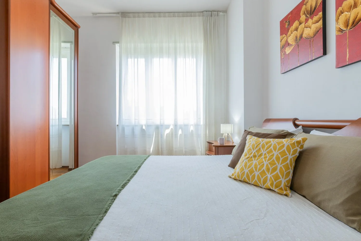 A bedroom with a neatly made bed, patterned cushions, and a bright curtained window.