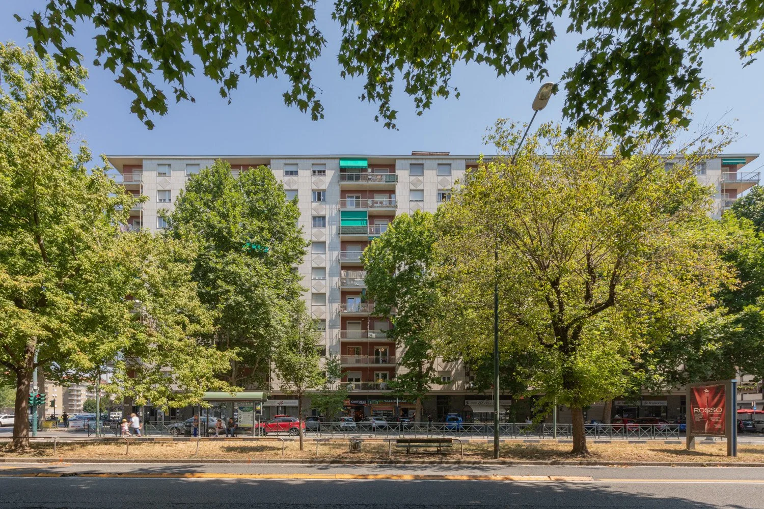 Apartment buildings line a tree-shaded street with storefronts at ground level.