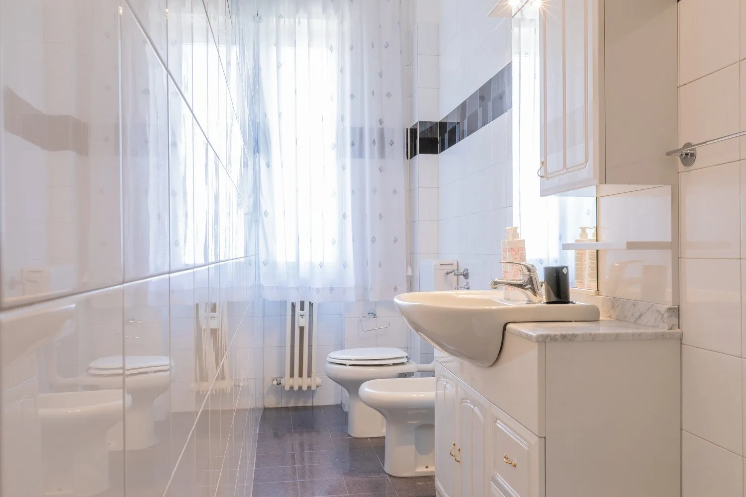 A bright tiled bathroom with a sink, toilet, and a sheer-curtained window.