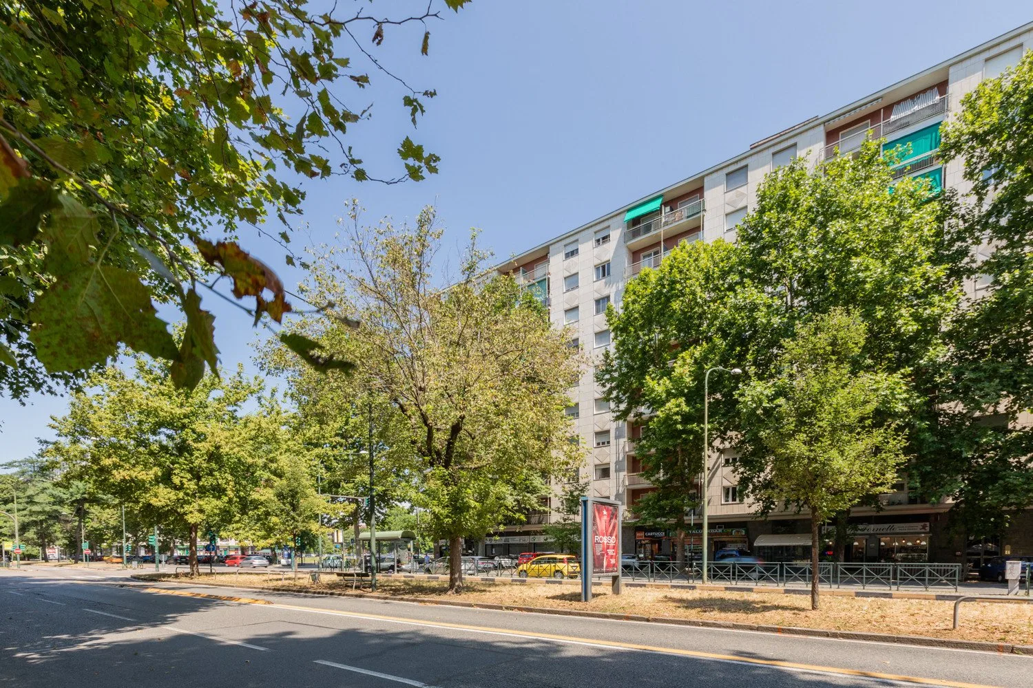 A tree-lined street runs beside a multistory building with ground-floor shops.