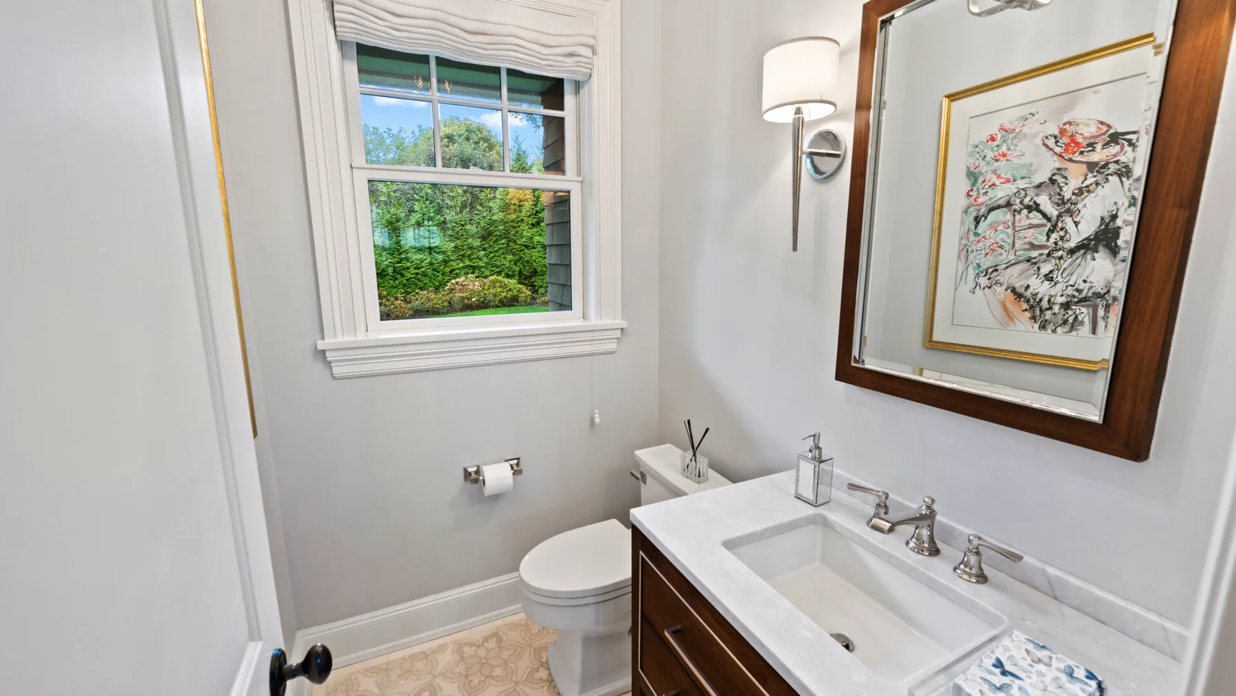 Un tocador de baño y un inodoro están bajo una ventana junto a una obra enmarcada.