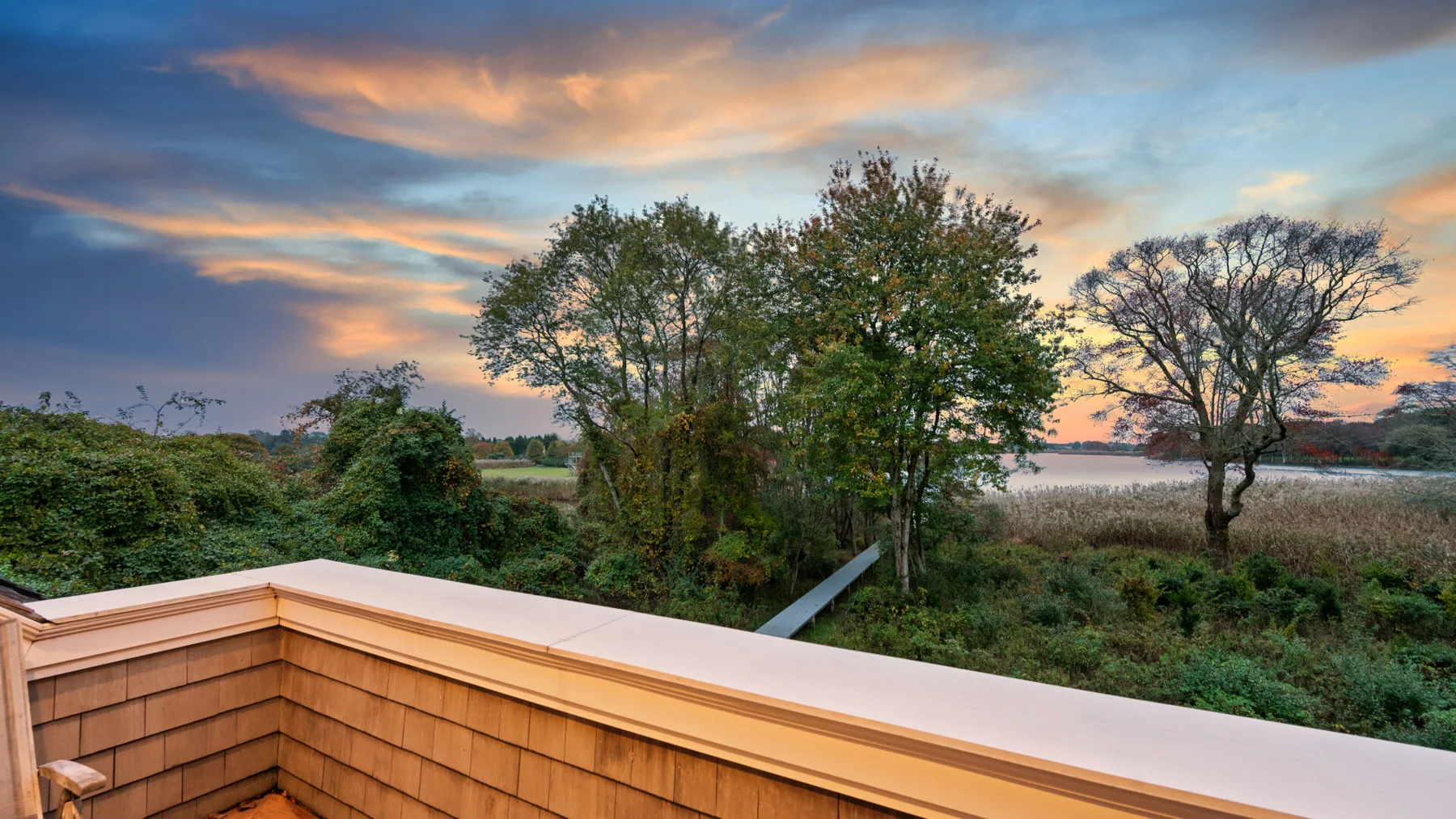 Una terraza de madera mira hacia árboles y marismas bajo un cielo colorido al atardecer.
