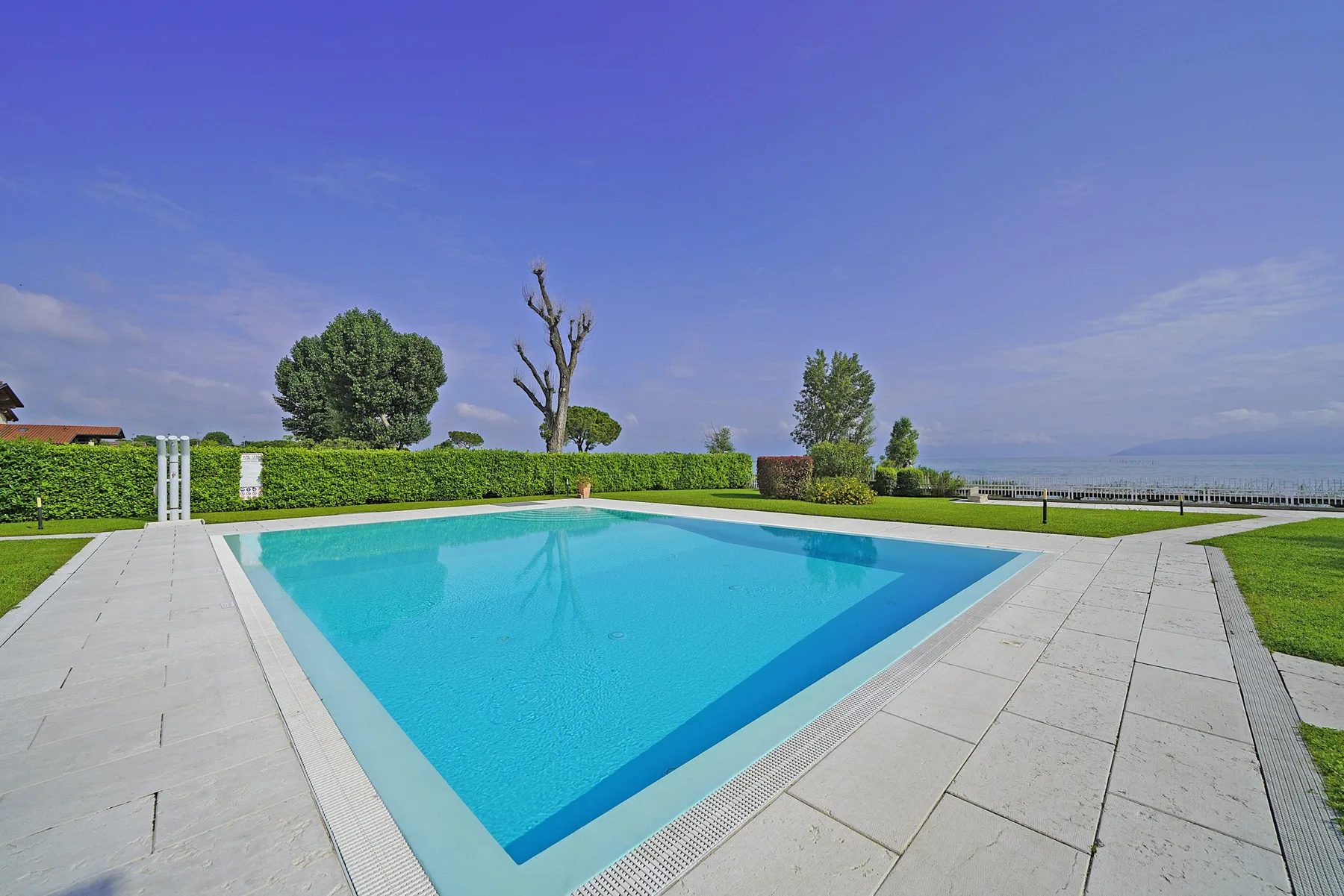 Una piscina al aire libre está rodeada por una terraza pavimentada y césped verde.