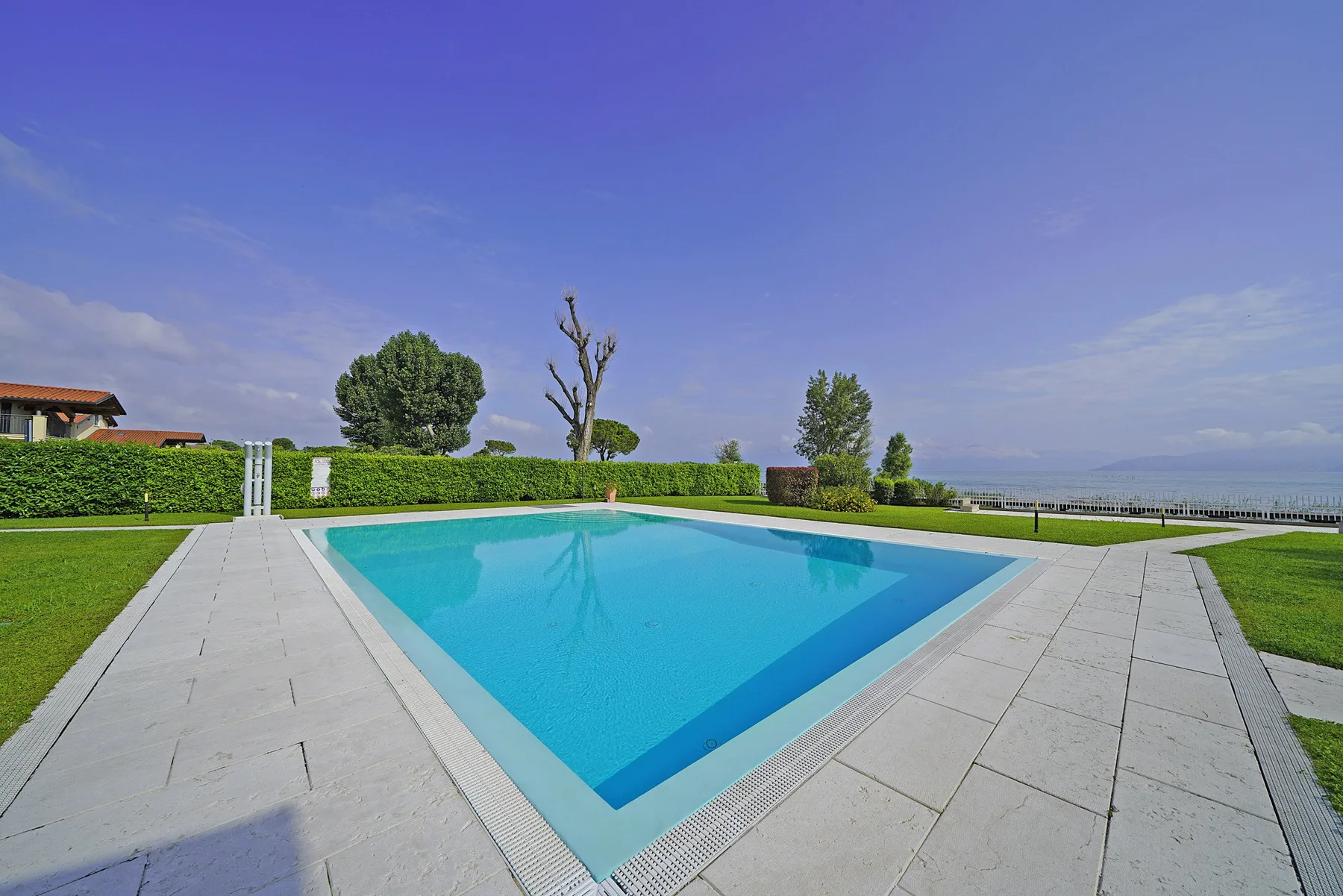 Una piscina al aire libre está rodeada por una terraza pavimentada y césped verde.