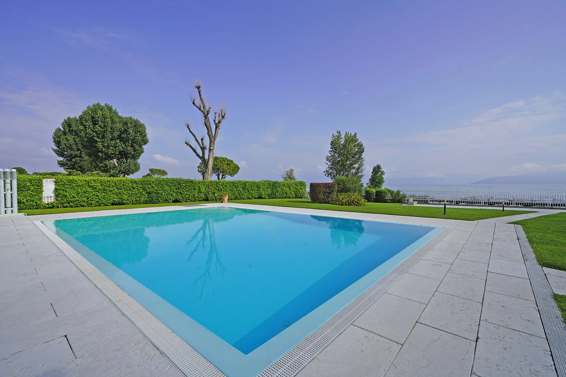 Una piscina al aire libre está rodeada de pavimento y setos verdes.