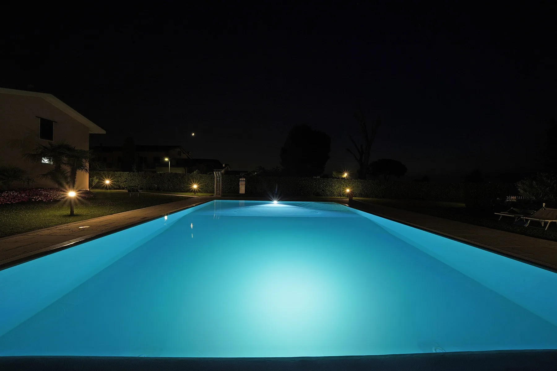 Una piscina exterior iluminada por la noche, rodeada de jardines.
