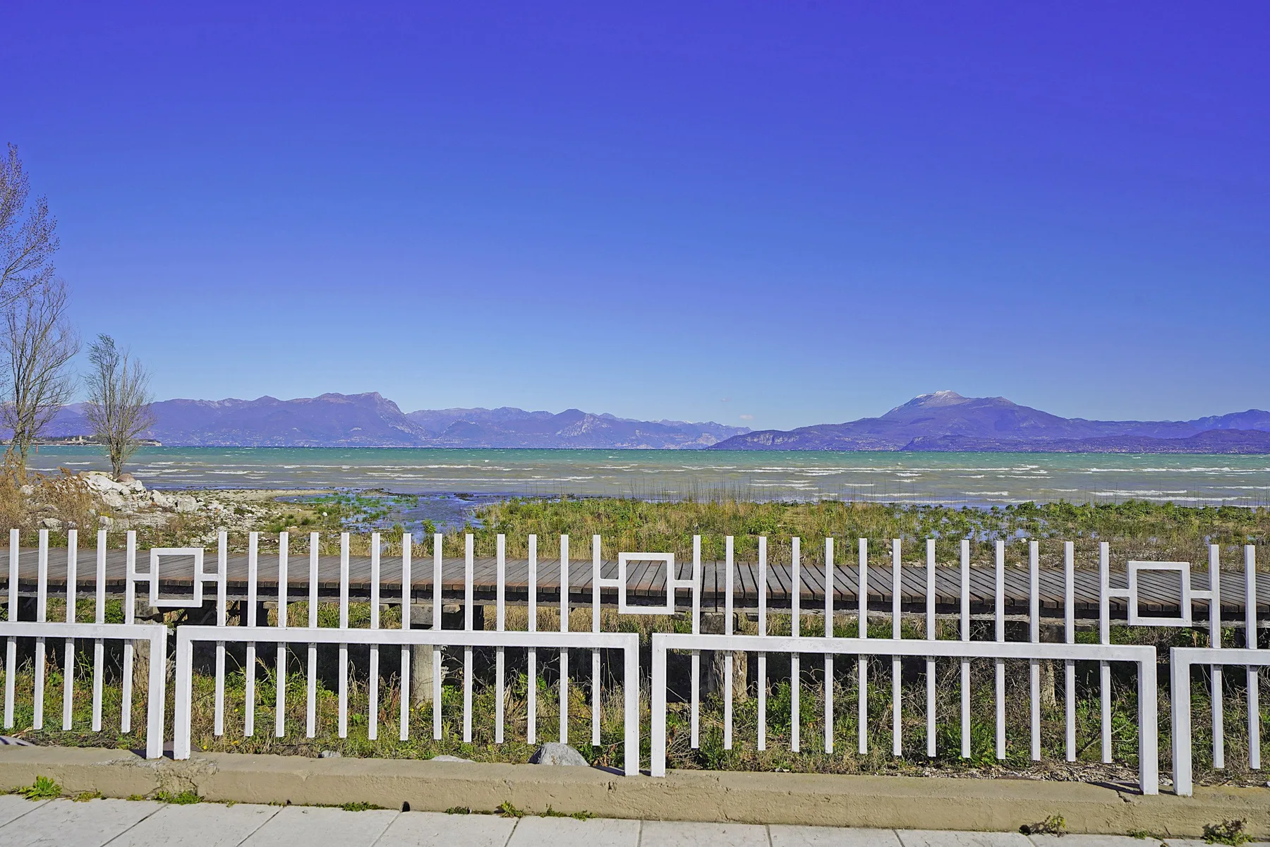 Una cerca blanca bordea una vista del lago y las montañas bajo un cielo azul despejado.