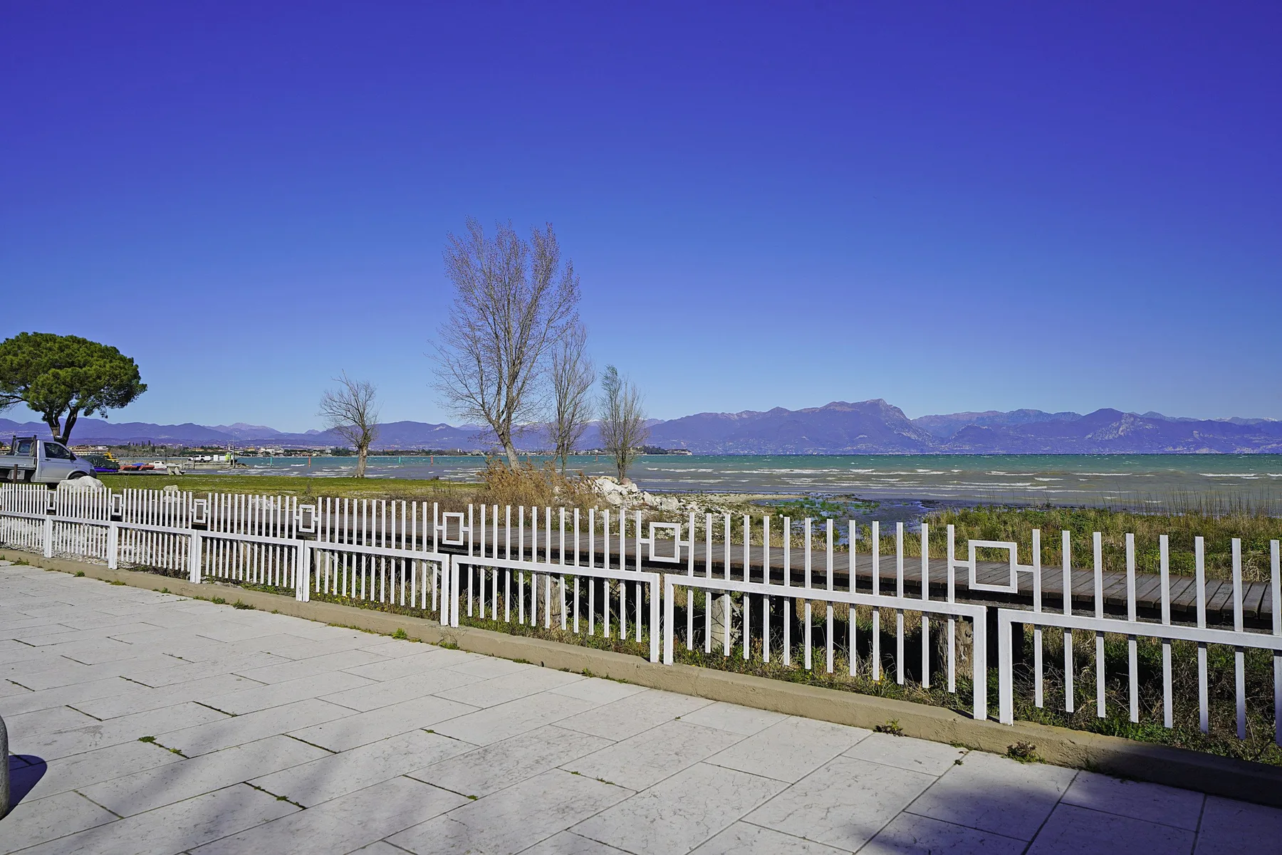 Una terraza pavimentada da a una orilla cercada, aguas azules y montañas lejanas.