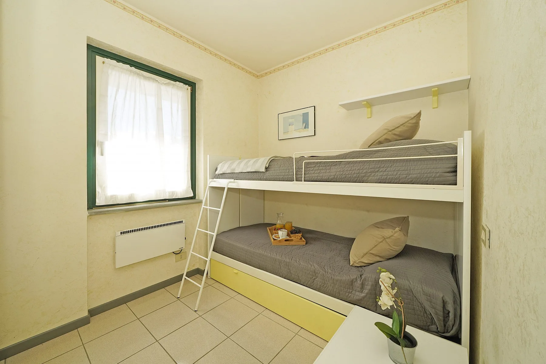 Un dormitorio con una litera blanca, ropa de cama gris y una ventana.