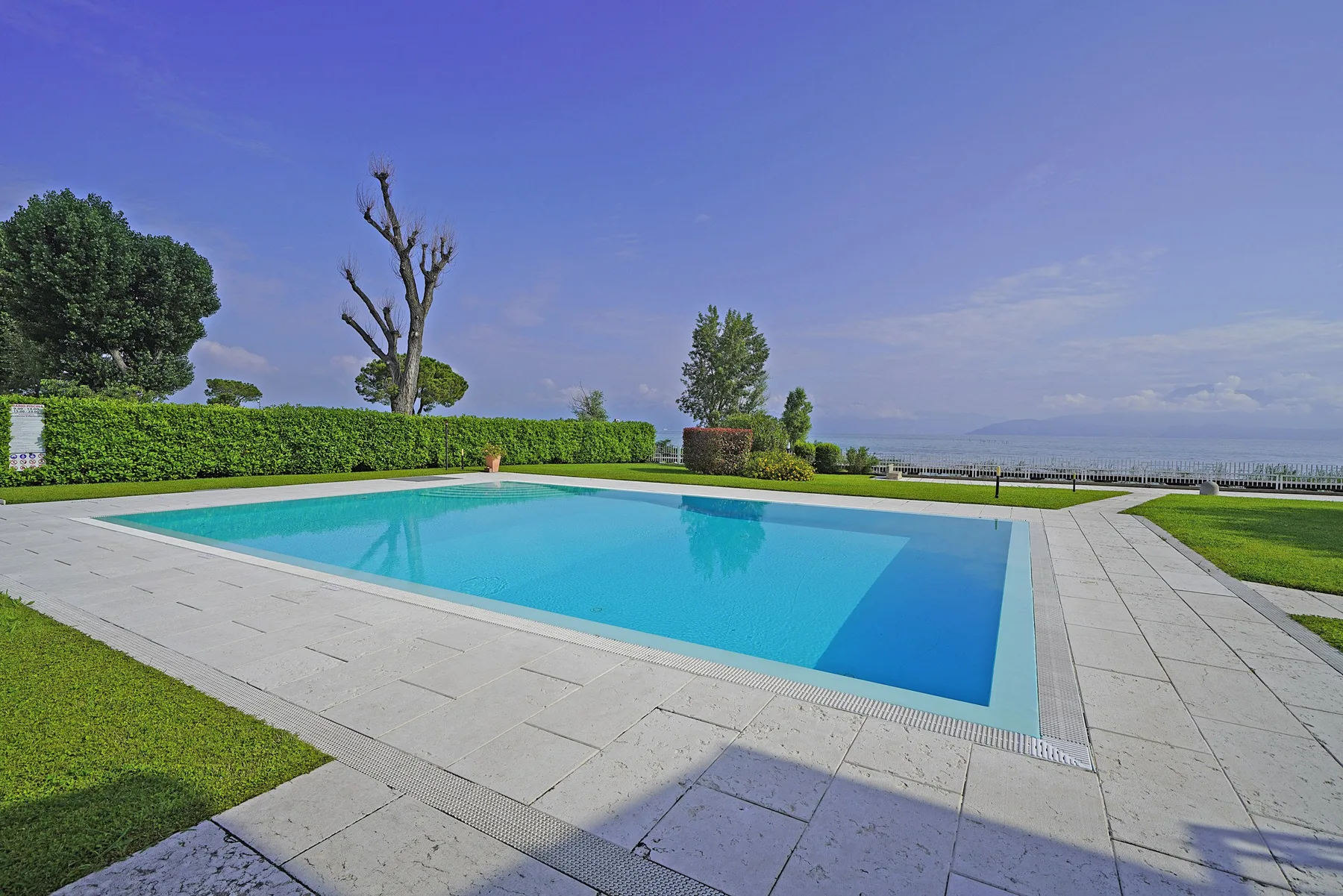 Una piscina al aire libre está rodeada por una terraza pavimentada y césped verde.