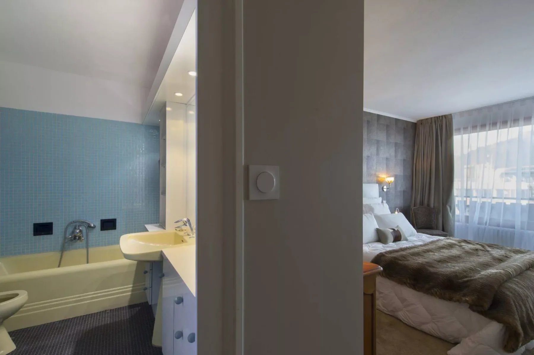 A bedroom with a bed and large curtained windows beside a bathroom with a bathtub.