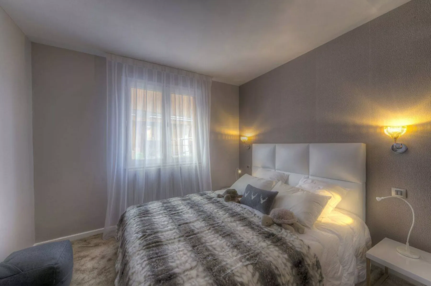 A bedroom with a bed, bedside lighting, and a window with sheer curtains.