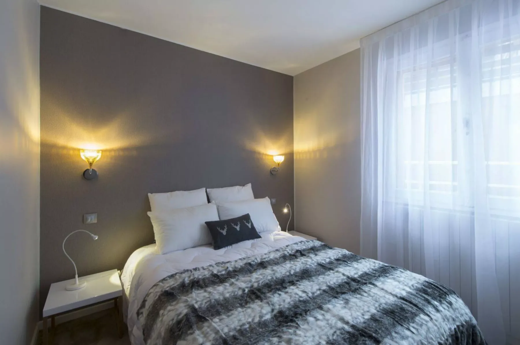 A bedroom with a neatly made bed beside a large curtained window.