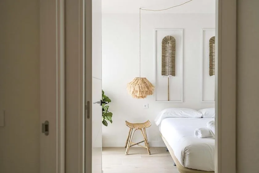 Un dormitorio luminoso con una cama blanca, una lámpara colgante tejida y un pequeño taburete de madera.