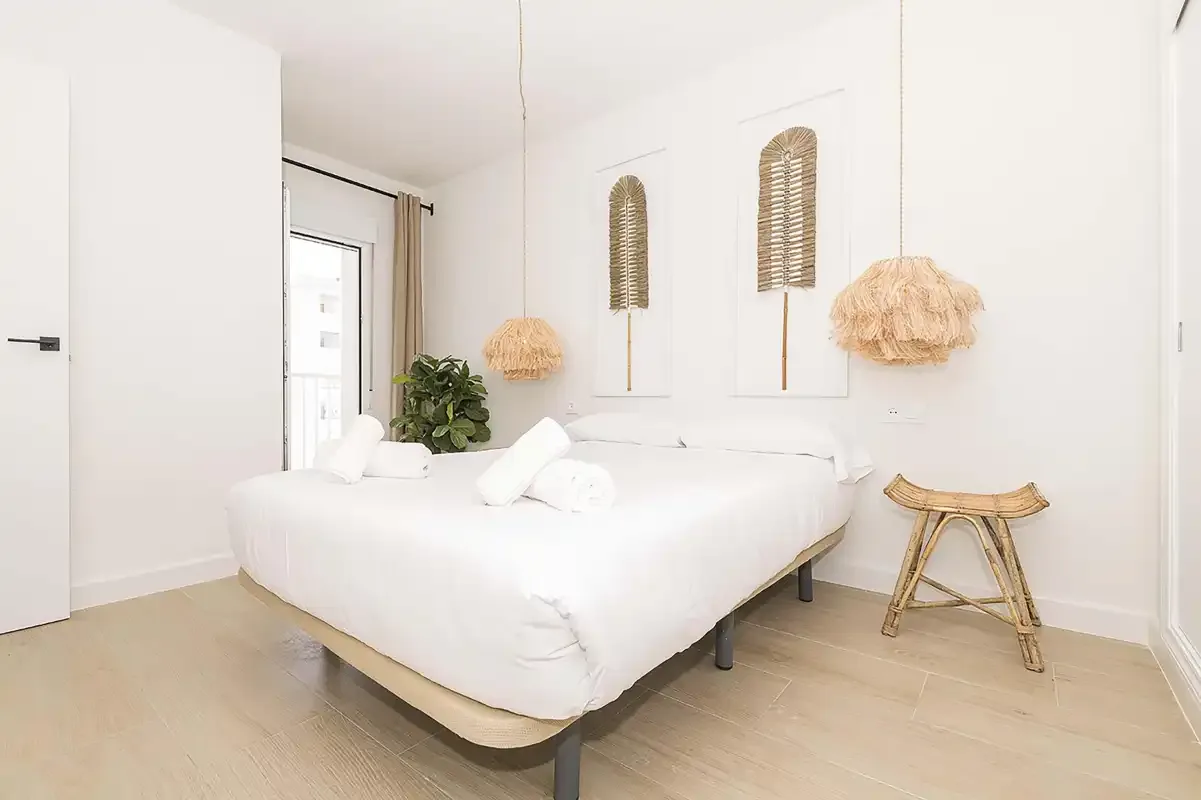 Un dormitorio luminoso con ropa de cama blanca, decoración tejida en la pared y un pequeño taburete de madera.
