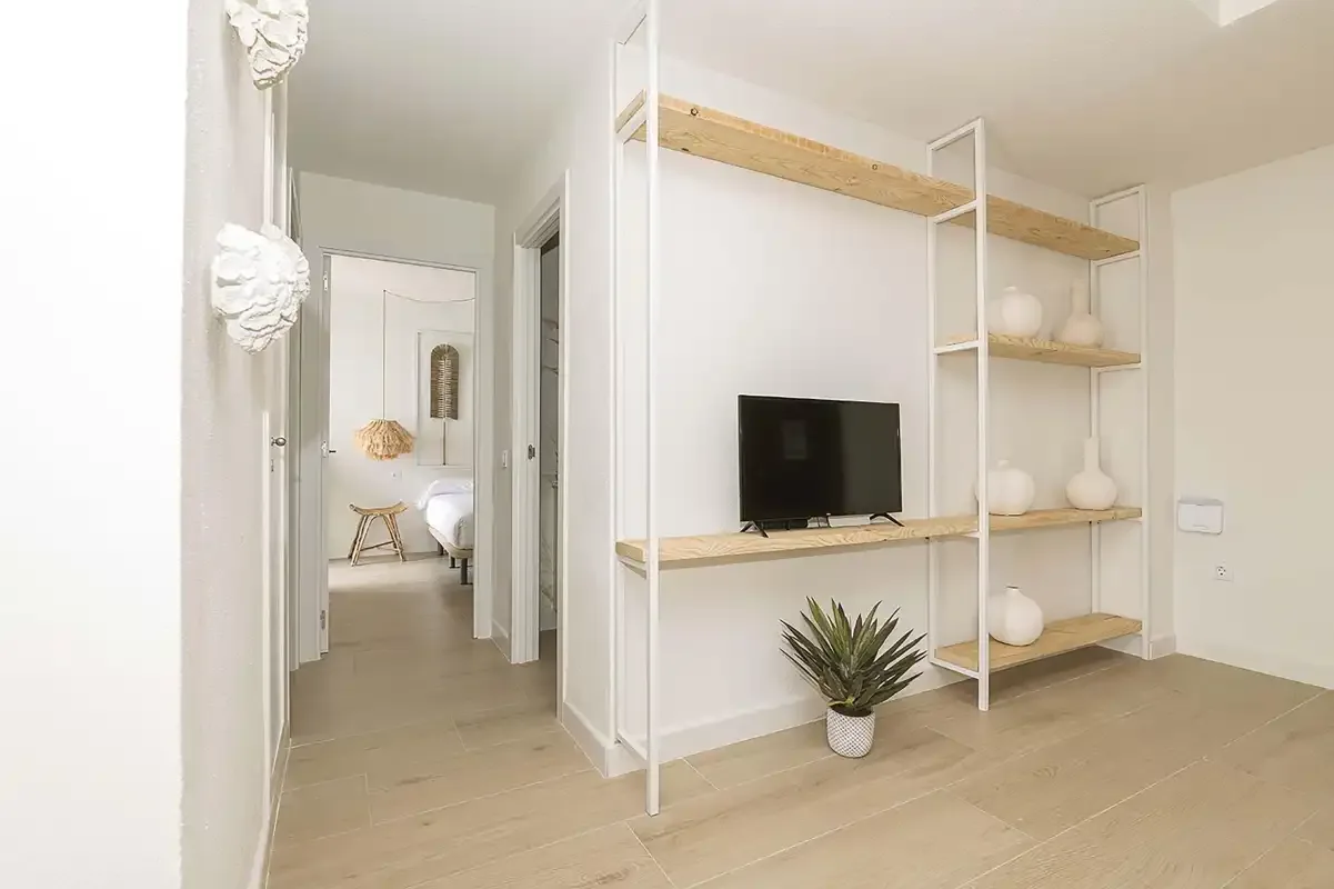 Un pasillo interior luminoso da a un dormitorio, con estanterías abiertas y un televisor.