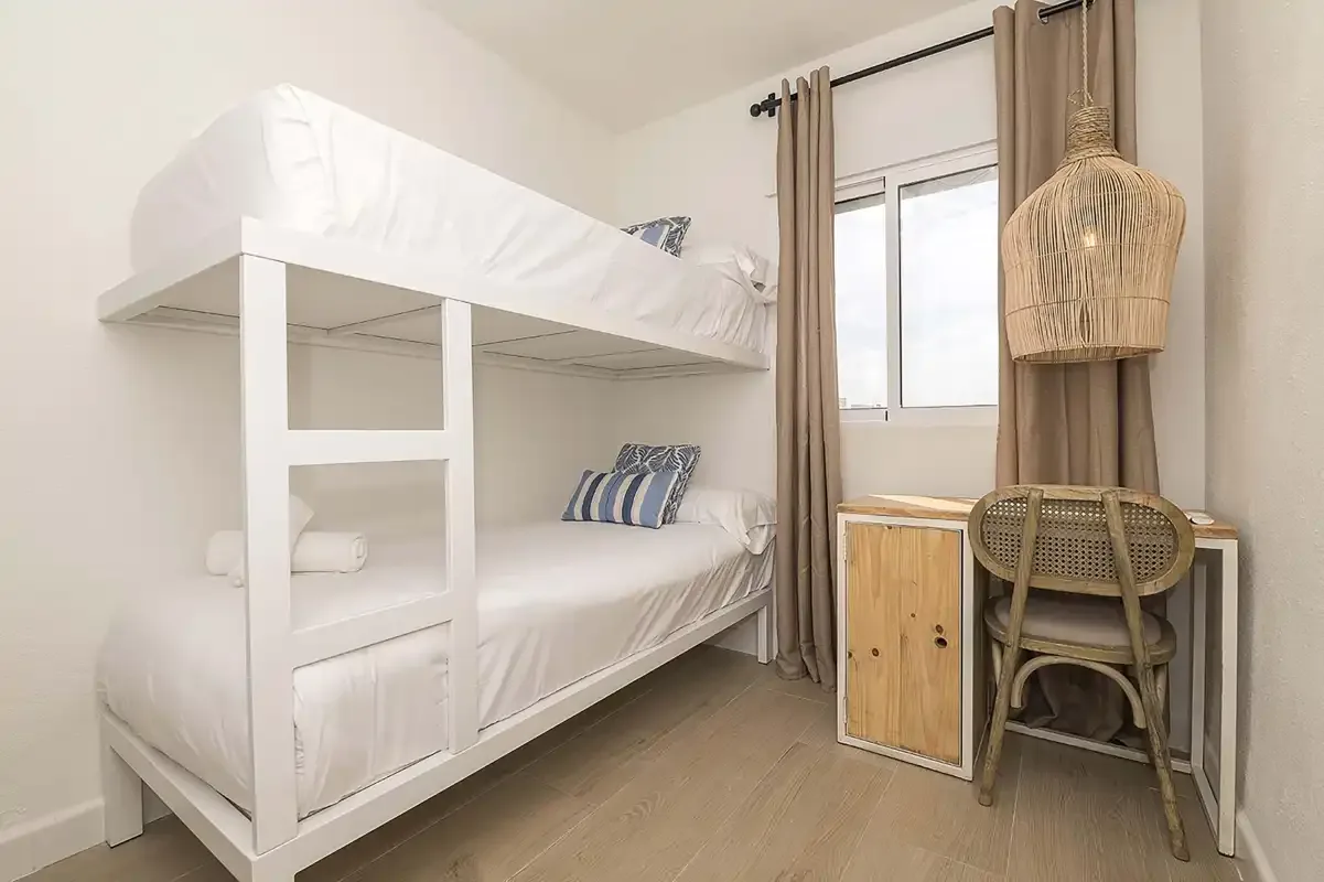 Un dormitorio luminoso con literas blancas y un escritorio junto a la ventana.