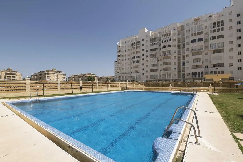 An outdoor swimming pool sits beside a paved deck, with apartment buildings in the background.