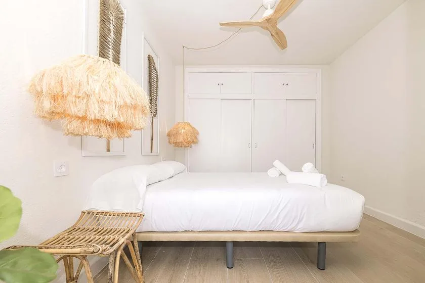 Un dormitorio luminoso con una cama blanca, asientos de fibras tejidas y un ventilador de techo.