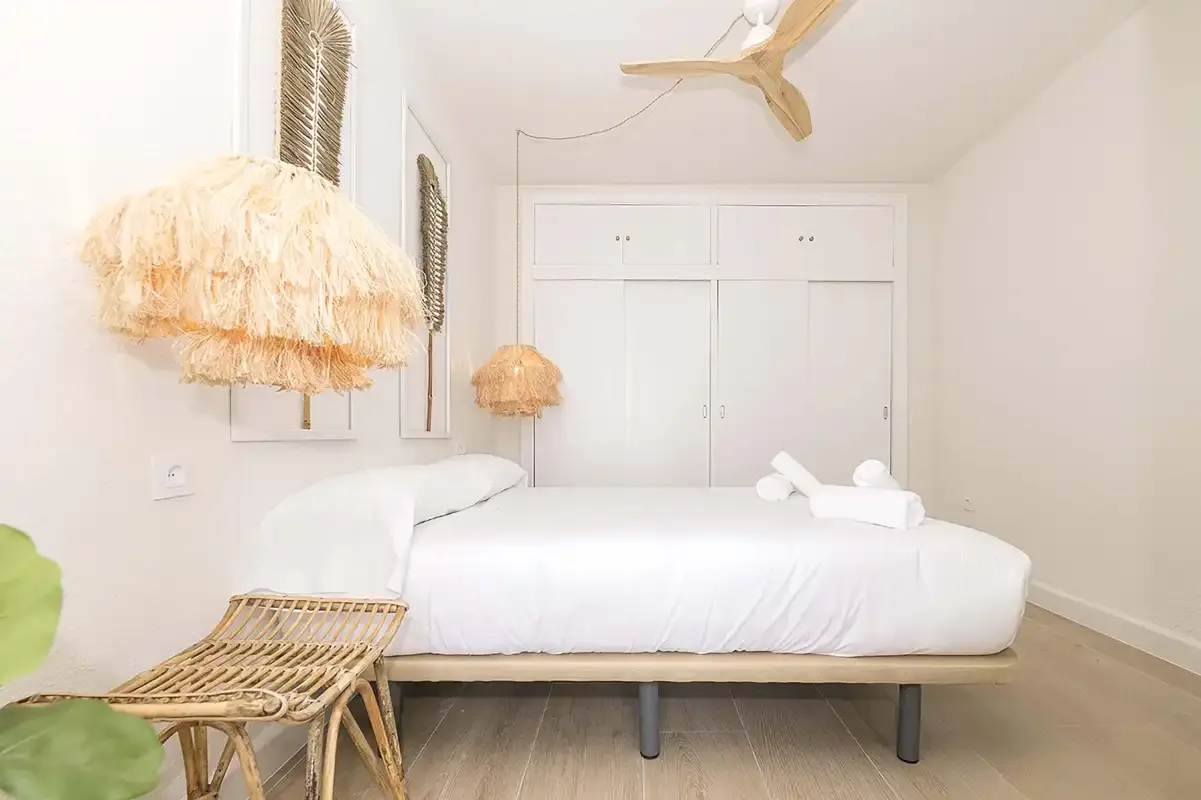 Un dormitorio luminoso con una cama blanca, asientos de mimbre y un ventilador de techo.