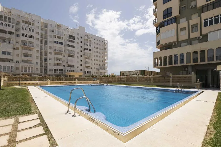 An outdoor swimming pool sits between multi-story buildings under a partly cloudy sky.