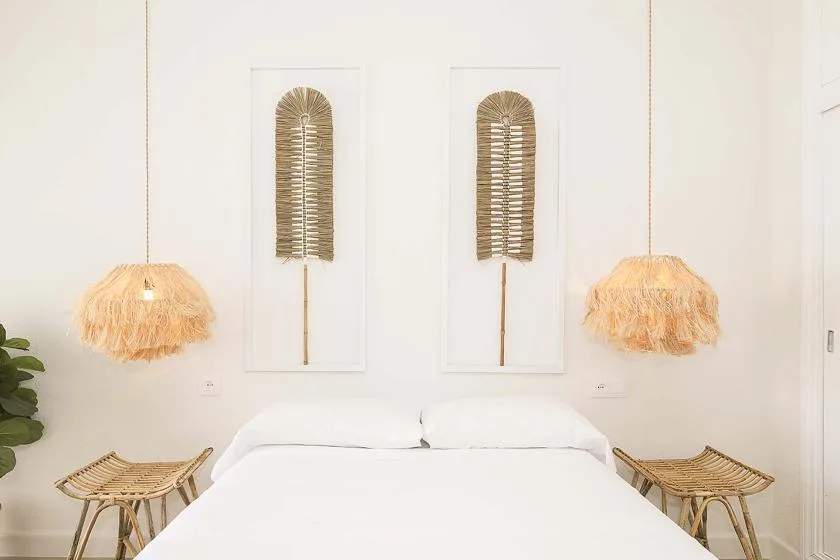 Un dormitorio luminoso con una cama blanca, arte tejido en la pared y lámparas colgantes.