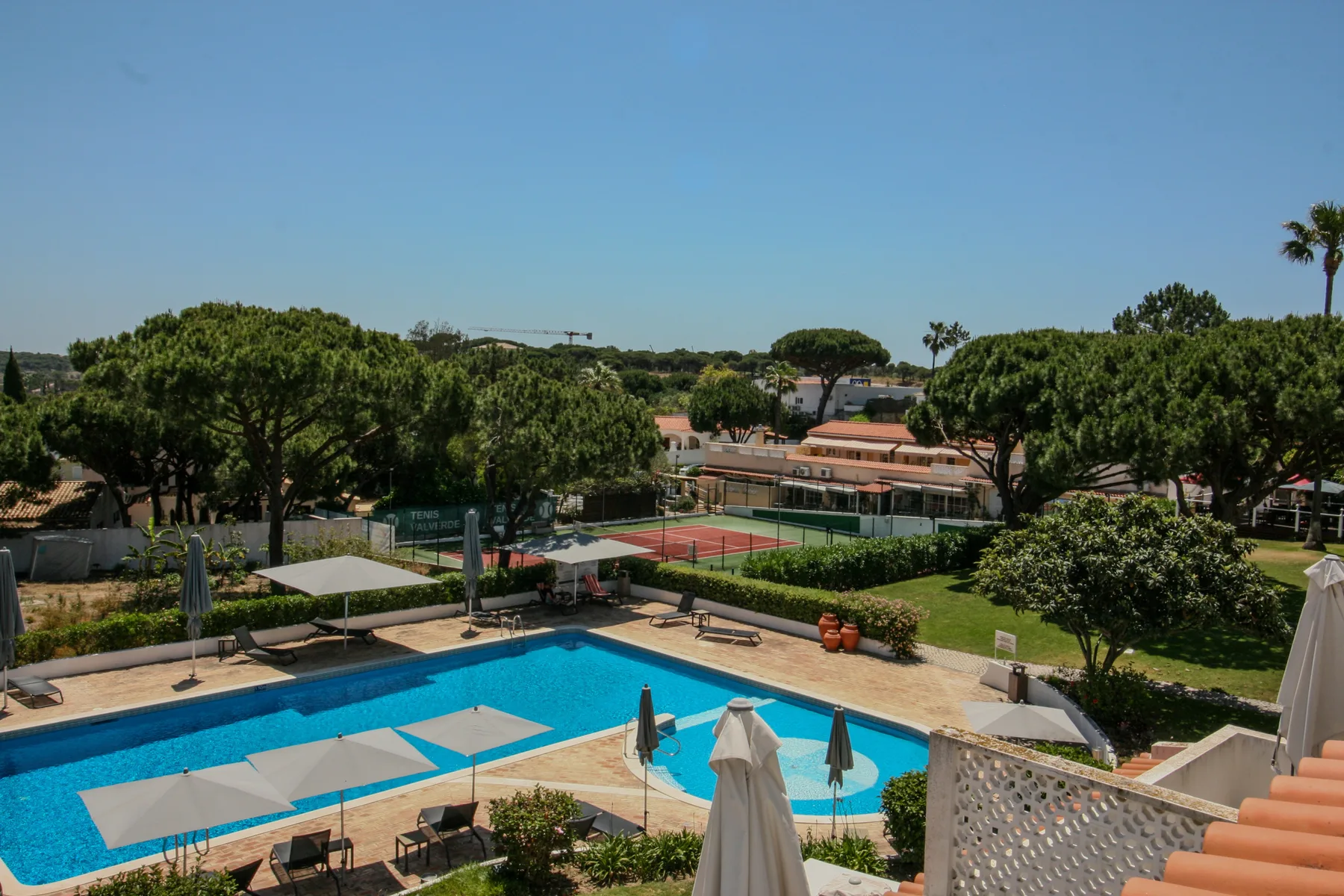 Una piscina al aire libre y una terraza pavimentada dan a jardines y edificios cercanos.