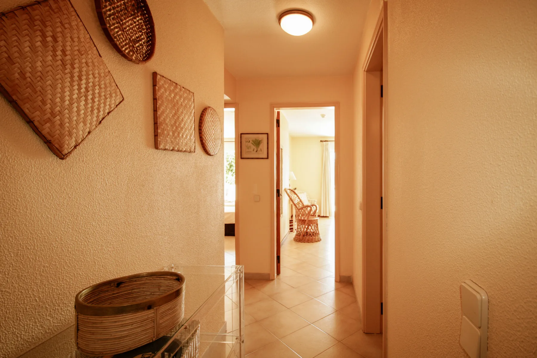 Un pasillo de tonos cálidos conduce a una habitación luminosa con una silla tejida.
